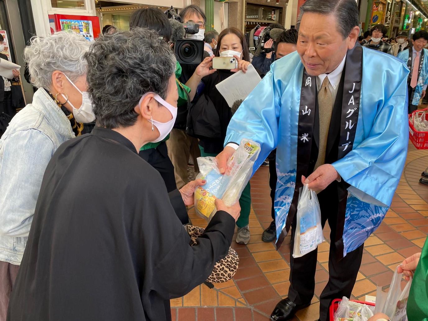 青江会長から「きぬむすめ」「にこまる」を受け取る商店街利用者の内容を表示